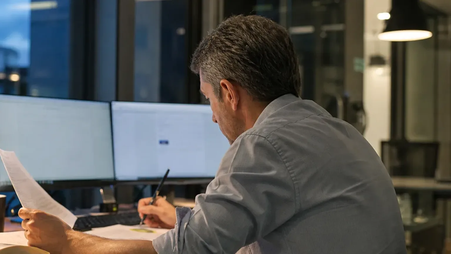 Operations analyst reading a printed vendor contract beside two monitors